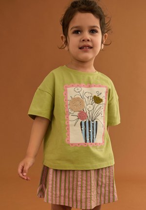 Junges Mädchen trägt ein grünes T-Shirt mit floralem Vasenmotiv und gestreiftem Rock, steht vor einem einfarbigen braunen Hintergrund.