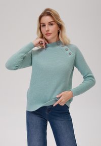 Pulover ribbed de culoare verde deschis, cu guler �înalt și nasturi decorativi pe umăr. Asortat cu blugi din denim albastru închis.