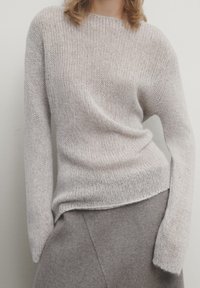Femme portant un pull en maille gris clair à manches longues et une jupe en laine grise, debout devant un fond uni pâle.