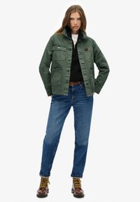 Groene katoenen jas met een kraagontwerp, meerdere voorzakken, gecombineerd met blauwe denim jeans en bruine veterschoenen.