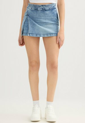 Mujer con minifalda de mezclilla azul con una abertura lateral, combinada con calcetines blancos hasta el tobillo y zapatillas blancas, de pie frente a un fondo sencillo.