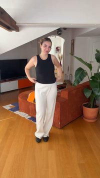 Νεαρή γυναίκα ρυθμίζει ένα μαύρο τοπ, φοράει λευκό παντελόνι και στέκεται σε ένα σαλόνι με καφέ καναπέ, μεγάλο φυτό και τηλεόραση στο παρασκήνιο.
