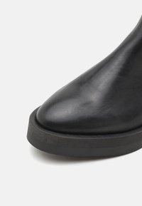 Botte noire en cuir jusqu'à la cheville avec un bout pointu, une texture lisse et une semelle noire épaisse et robuste au fini mat.