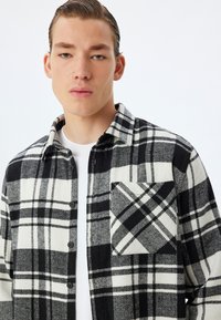 Camicia di flanella a scacchi bianco e nero con motivo a spina di pesce, dotata di design con bottoni e taschino sul lato sinistro.