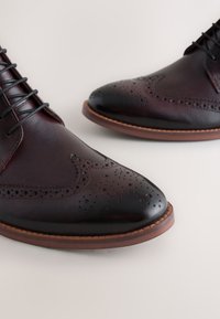 Braune Lederschuhe mit glatter Oberfläche, verziert mit Brogue-Details, spitzer Zehe und Schnürverschluss. Gummisohle mit dezenter brauner Umrandung.