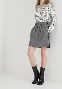 Mujer llevando una blusa blanca con lunares negros, una falda de cuadros de cintura alta y unas botas de tacón negro brillantes hasta la mitad de la pantorrilla.