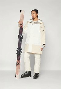 Completo da sci con giacca impermeabile bicolore beige e bianca, pantaloni coordinati e scarponi da sci neri. Persona che tiene degli sci marroni.