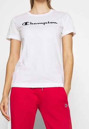 Frau trägt weißes Champion-T-Shirt mit schwarzem Logo und rote Hose mit weißen Streifen und einem Logo am linken Oberschenkel.