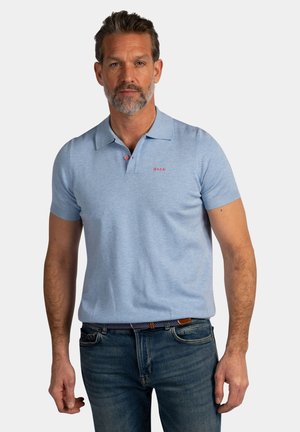 Man met baard, gekleed in een lichtblauw poloshirt met korte mouwen en donkerblauwe spijkerbroek, staand tegen een effen achtergrond.
