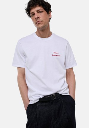 Homme aux cheveux bouclés et foncés, portant un T-shirt blanc avec un texte rouge « Daron d'exception » et un pantalon sombre avec une ceinture noire, mains dans les poches.