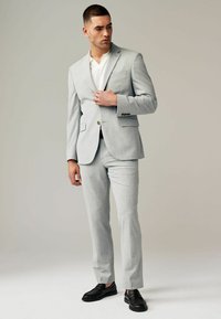 Traje gris claro con una chaqueta y pantalones a medida. Presenta una textura sutil, dos bolsillos delanteros y zapatos negros de mocasín.