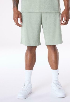 Mann trägt hellgrüne Shorts, weiße Socken und weiße Sneakers und steht mit entspannten Händen an den Seiten.