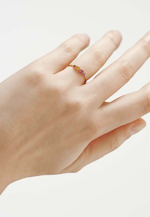 Mano con un anillo de oro con pequeñas piedras preciosas naranjas y moradas en el dedo anular sobre un fondo blanco.