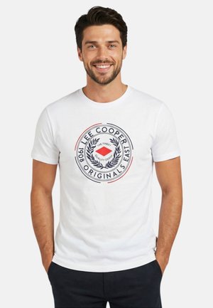 Homme souriant aux cheveux foncés portant un t-shirt blanc avec un logo circulaire Lee Cooper Originals et un pantalon foncé, les mains dans les poches.