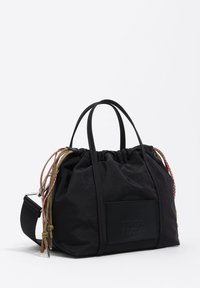 Bolso de tela negro con cierre de cordón, asas cortas y una correa ajustable. Presenta detalles texturizados y un parche con el logo.