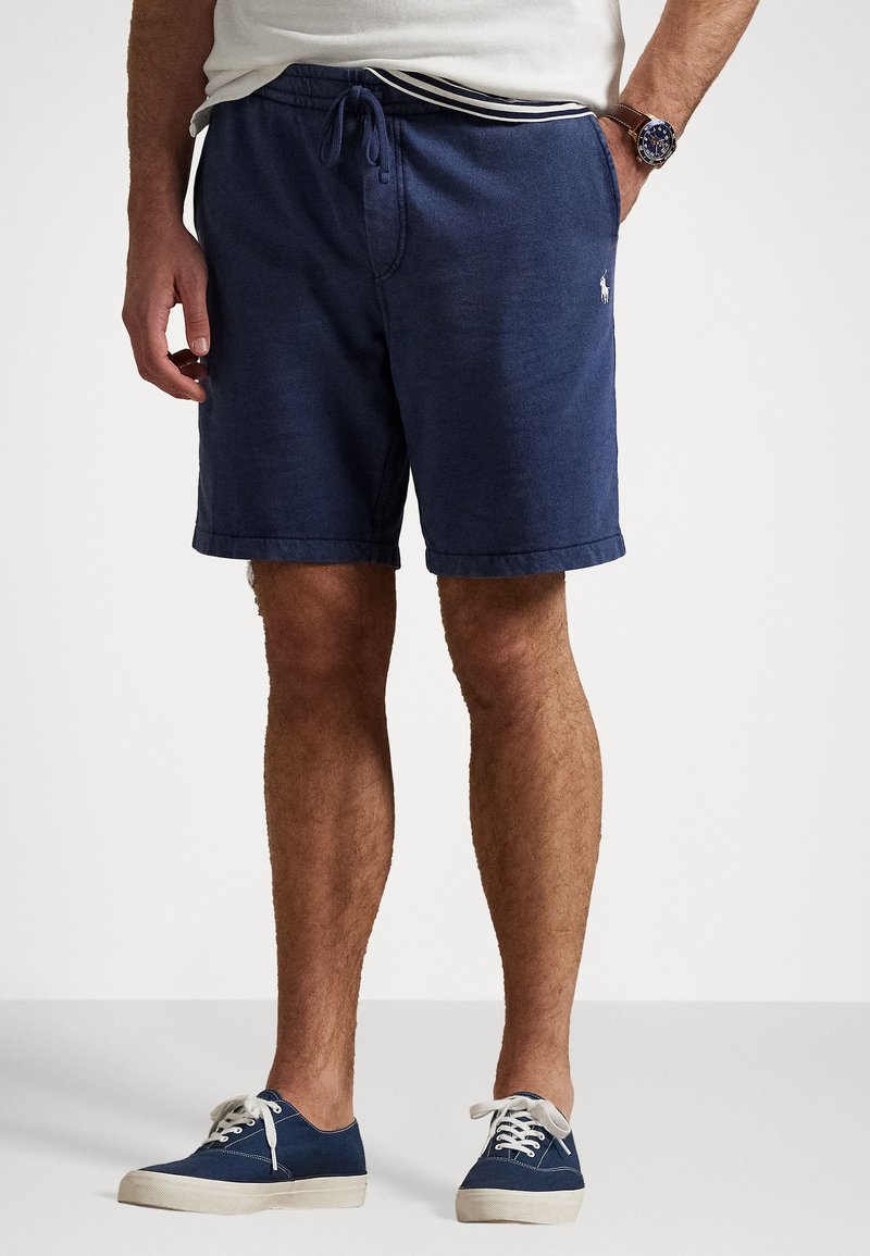 Shorts bleu marine en tissu respirant, avec un cordon de serrage à la taille et un ourlet retroussé. Un petit logo est visible sur la cuisse gauche.