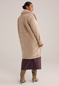 Manteau en peluche beige avec un col haut, des manches longues et une coupe décontractée, associé à une jupe bordeaux foncé et des bottes taupe à genoux.