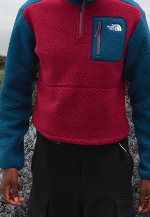 Kurtka z polaru z bordowym korpusem i zielononiebieskimi rękawami, z zielononiebieską kieszenią na piersi na suwak z logo The North Face.