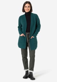 Teal knit cardigan with open front, ribbed texture, and two front pockets, paired with dark pants and black ankle boots.