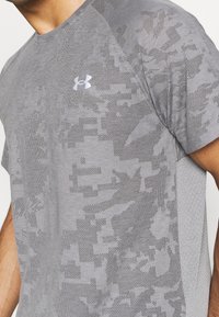 Camiseta atlética gris con un patrón de camuflaje texturizado. Logo de Under Armour en plateado en el pecho. Mangas cortas y tejido transpirable.