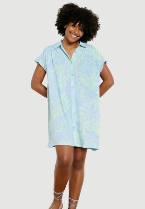Lächelnde Frau mit lockigem Haar, die ein lockeres, hellblaues Kleid mit kurzen Ärmeln, Knöpfen und grünem Blättermuster trägt und mit den Händen hinter dem Rücken steht.