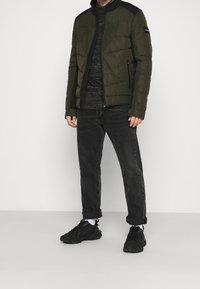 Olivgrön vadderad jacka med svarta detaljer, dragkedja, två sidofickor, bärs över en mörk skjorta och svarta jeans, matchad med svarta sneakers.