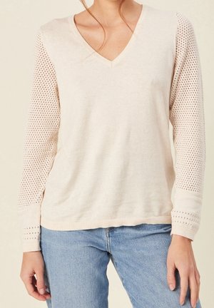Femme portant un pull léger beige à col en V avec des manches longues perforées et un jean bleu clair, sur un fond uni.