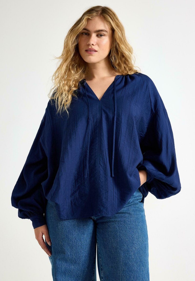 Marinblå, oversized blus med v-ringning, långa puffärmar och strukturerad tyg. Matchad med blå denimjeans.