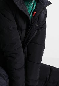 Czarna pikowana kurtka z suwakiem i zatrzaskami częściowo rozpięta, pod nią zielony element ubrania z okrągłym srebrnym pierścieniem jako uchwytem.