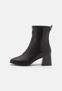 Bottine noire en cuir lisse, avec un bout pointu, une fermeture éclair et un talon épais avec un accent métallique.