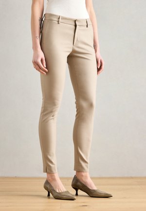Femme portant un pantalon beige slim et des chaussures marron à talons bas, debout sur un sol en bois devant un mur clair uni.