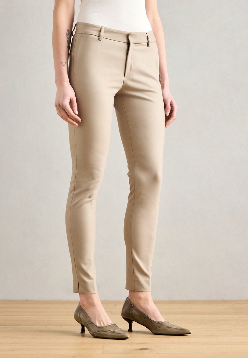 Femme portant un pantalon beige slim et des chaussures marron à talons bas, debout sur un sol en bois devant un mur clair uni.