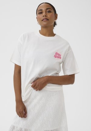 Kvinde iført hvid "Sunset Chaser" t-shirt og tekstureret hvid nederdel, stående mod en ensfarvet lys baggrund.