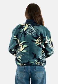 Chaqueta de forro polar floral en verde azulado con un cuello marino a contraste. Presenta un gran estampado floral en tonos crema y oscuros, corte relajado y dobladillo acanalado.