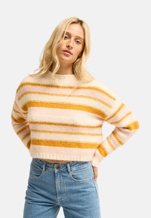 Femme aux cheveux blonds portant un pull court rayé dans des tons crème, jaune et rose, et un jean bleu taille haute, posant les mains sur les hanches.