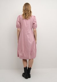 Rosa klänning med korta puffärmar, figursydd bodice och A-linjeformad kjol. Slät textur, knälång med blixtlås baktill. Svarta stövlar.