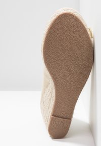 Chaussure beige à enfiler avec une tige en tissu texturé et une semelle en caoutchouc dotée d'une adhérence à motif. L'arrière présente un bord lisse et un petit logo.