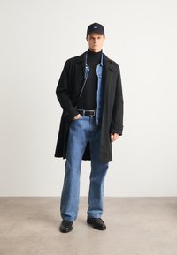 Un manteau trench noir porté sur un col roulé noir et une veste en denim superposée. Un jean large bleu clair et des chaussures noires complètent la tenue.