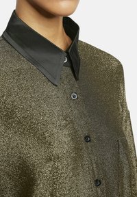 Vrouw in een glanzend gouden overhemdbloes met een zwarte puntkraag, waarbij de bovenste torso en nek zichtbaar zijn tegen een effen achtergrond.