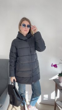 Casaco puffer preto com capuz, design acolchoado, duas bolsos frontais, combinado com jeans azul claro e mochila preta. Óculos de sol e sapatilhas rosas.