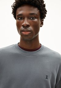 Sweatshirt gris avec un col rond et un petit logo bleu marine sur la poitrine. Texture douce avec une coupe décontractée, superposé à une chemise bordeaux.