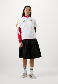 Άσπρο κοντομάνικο πουκάμισο με μαύρο λογότυπο και κόκκινες λεπτομέρειες, τοποθετημένο πάνω από ένα κόκκινο μακρυμάνικο πουκάμισο, συνδυασμένο με μια μαύρη πλισέ φούστα. Ασημένια αθλητικά παπούτσια.