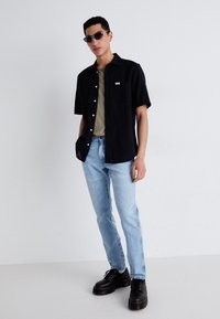 Camisa negra de manga corta con un bolsillo, combinada con una camiseta verde oliva debajo. Un pantalón vaquero azul claro y unos zapatos negros de suela gruesa completan el look.