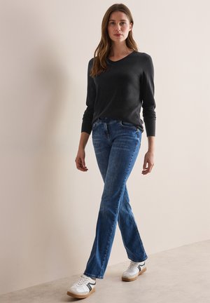 Mörkgrå V-ringad tröja i kombination med ljusblå utsvängda jeans och vita sneakers med svarta ränder, mot en neutral bakgrund.