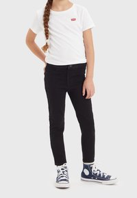 Camiseta blanca de manga corta con un logo rojo, combinada con jeans negros ajustados. Lleva zapatillas altas azules con acentos blancos.