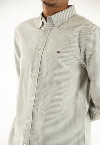 Camicia button-up grigia chiara con sottili righe bianche. Presenta un colletto a punta, maniche lunghe e un piccolo logo sul petto.
