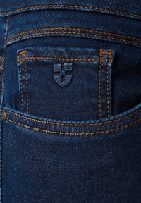 Jeans dżinsowe mają ciemnoniebieski kolor z pomarańczowym przeszyciem, małą tylną kieszeń oraz wytłoczone logo na szwie kieszeni.