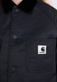 Schwarze Jacke mit glänzendem Finish, ausgestattet mit einem schwarzen Samtkragen und einer Fronttasche mit einem weißen Carhartt-Logo-Patch. Matte Knöpfe sind sichtbar.