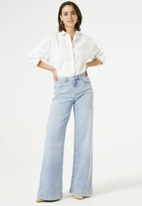 Chemise blanche à boutons avec manches retroussées, associée à un jean bleu clair à jambes larges. Le jean présente une taille haute et une finition délavée.
