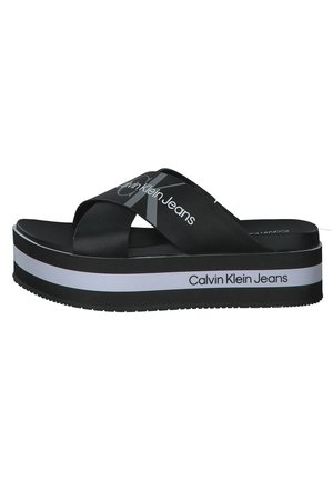 Czarna sandałka na platformie z krzyżującymi się paskami i białym napisem „Calvin Klein Jeans” na paskach i krawędzi podeszwy.